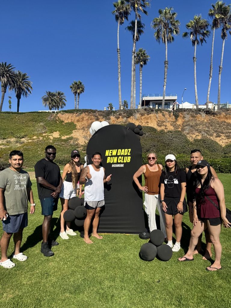 The New Dadz Run Club cookout crew posing around the NDRC backdrop on the grass, palm trees behind