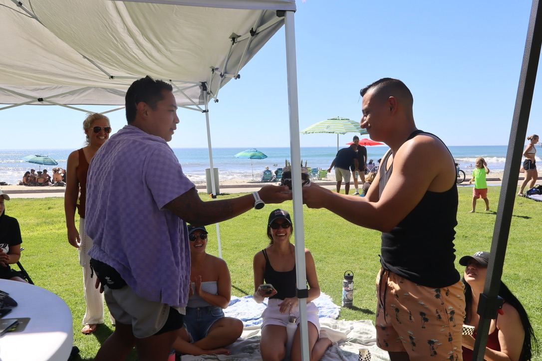 Two dads handing off food under the tent with the ocean behind